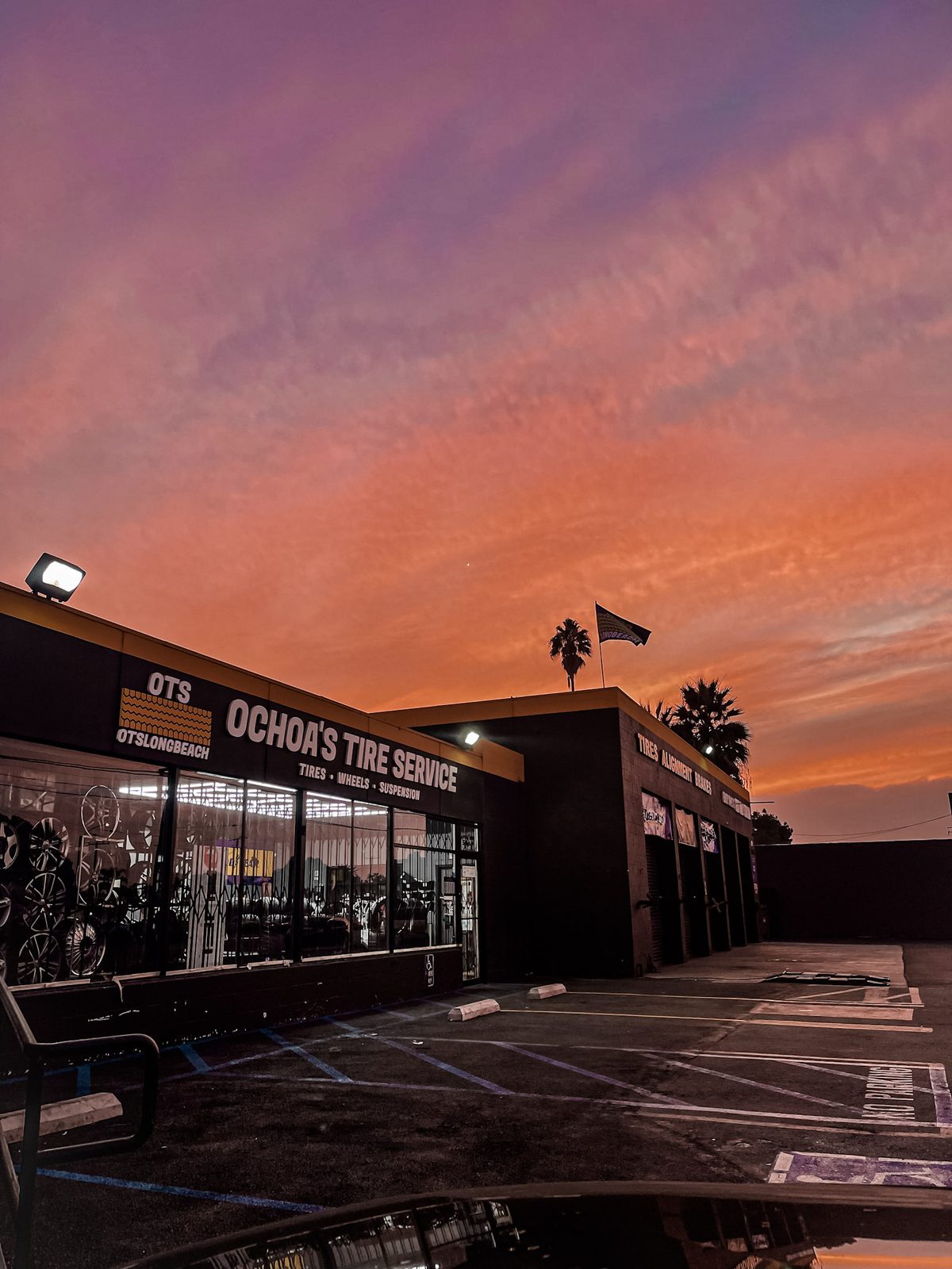 Ochoa's Tire Service storefront on Cherry Ave at sunset, with the OTS sign lit and a palm tree silhouetted against a pink and orange sky